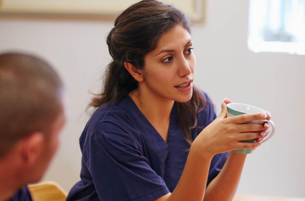 A female nurse in scrubs holding a cup of tea whilst in conversation