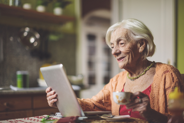 A senior woman using a tablet in her kitchen whilst holding a cup of tea in her left hand