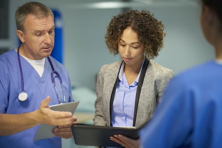 a mature hospital doctor is chatting with a team of fellow medical professionals , including a suited woman who could be a sales professional or administrator