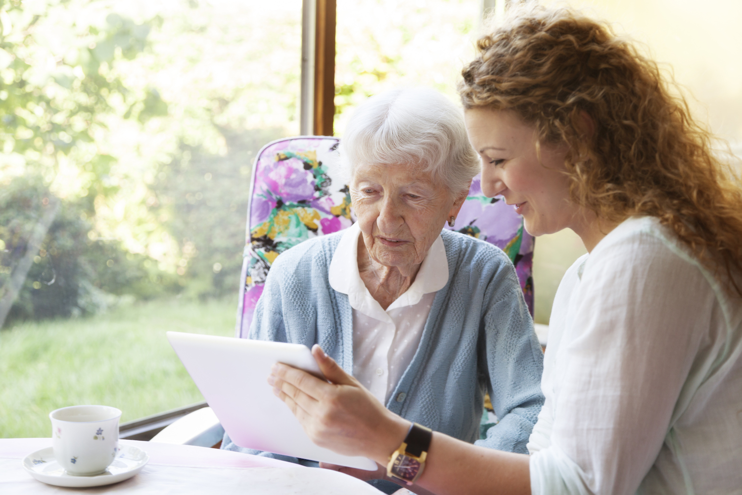 Young caregiver showing something on digital tablet to with 90 years old senior woman, sitting in nice conservatory