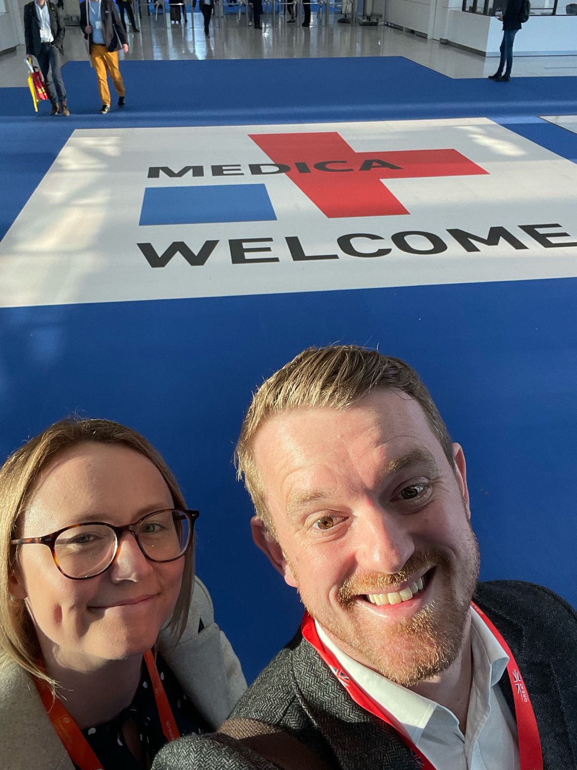 A woman and a man taking a selfie and smiling in front of a sign for MEDICA trade fair