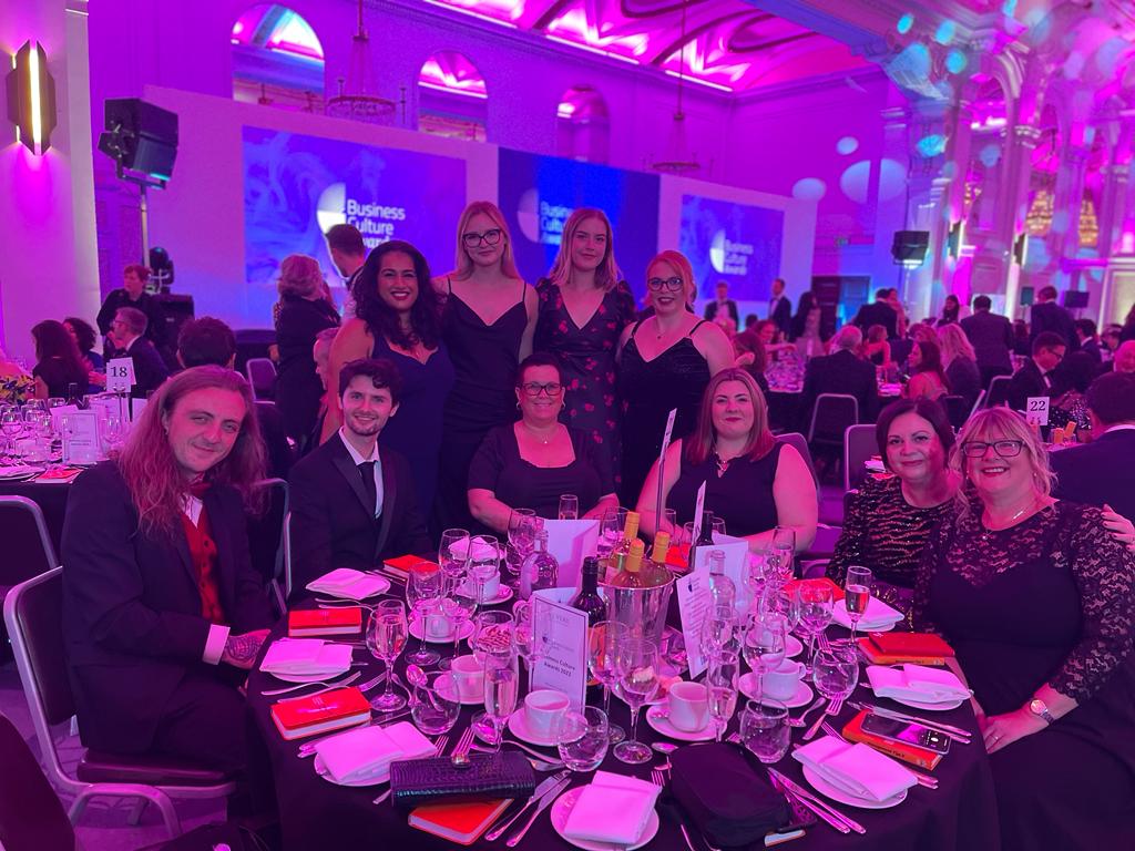 Group of people sat at a table with a row of people stood behind them. At an awards ceremony and dinner. All smiling