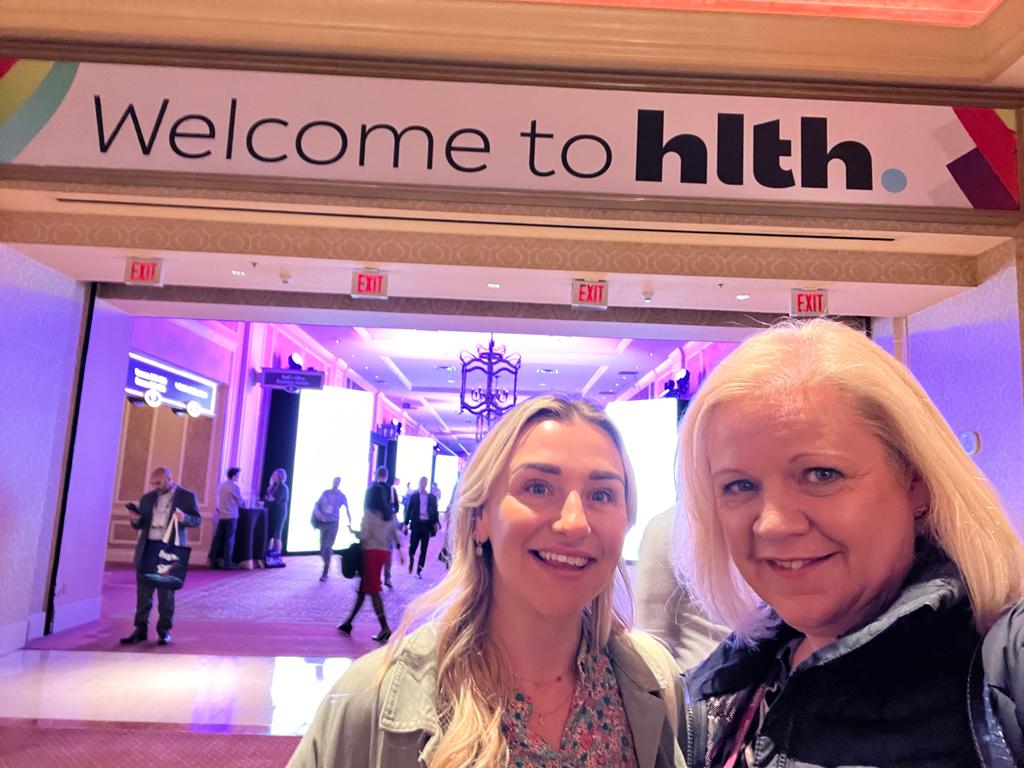 Selfie of women smiling in front of sign for HLTH conference